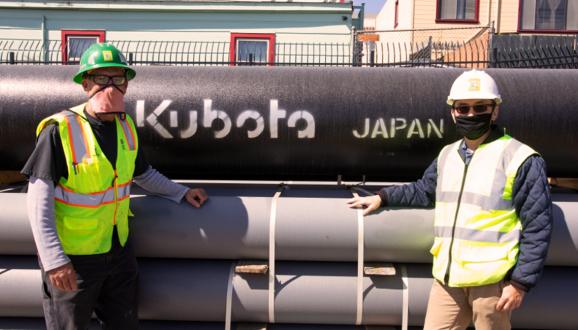 Two people standing in front of large pipes.